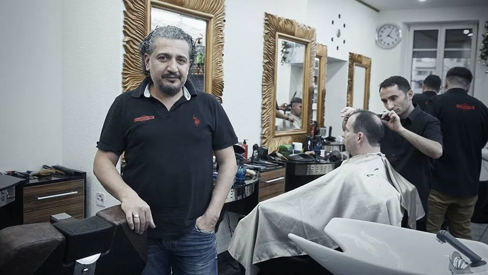 Ein Mitarbeiter im Salon, hinter ihm die Spiegel und ein Kunde im Stuhl